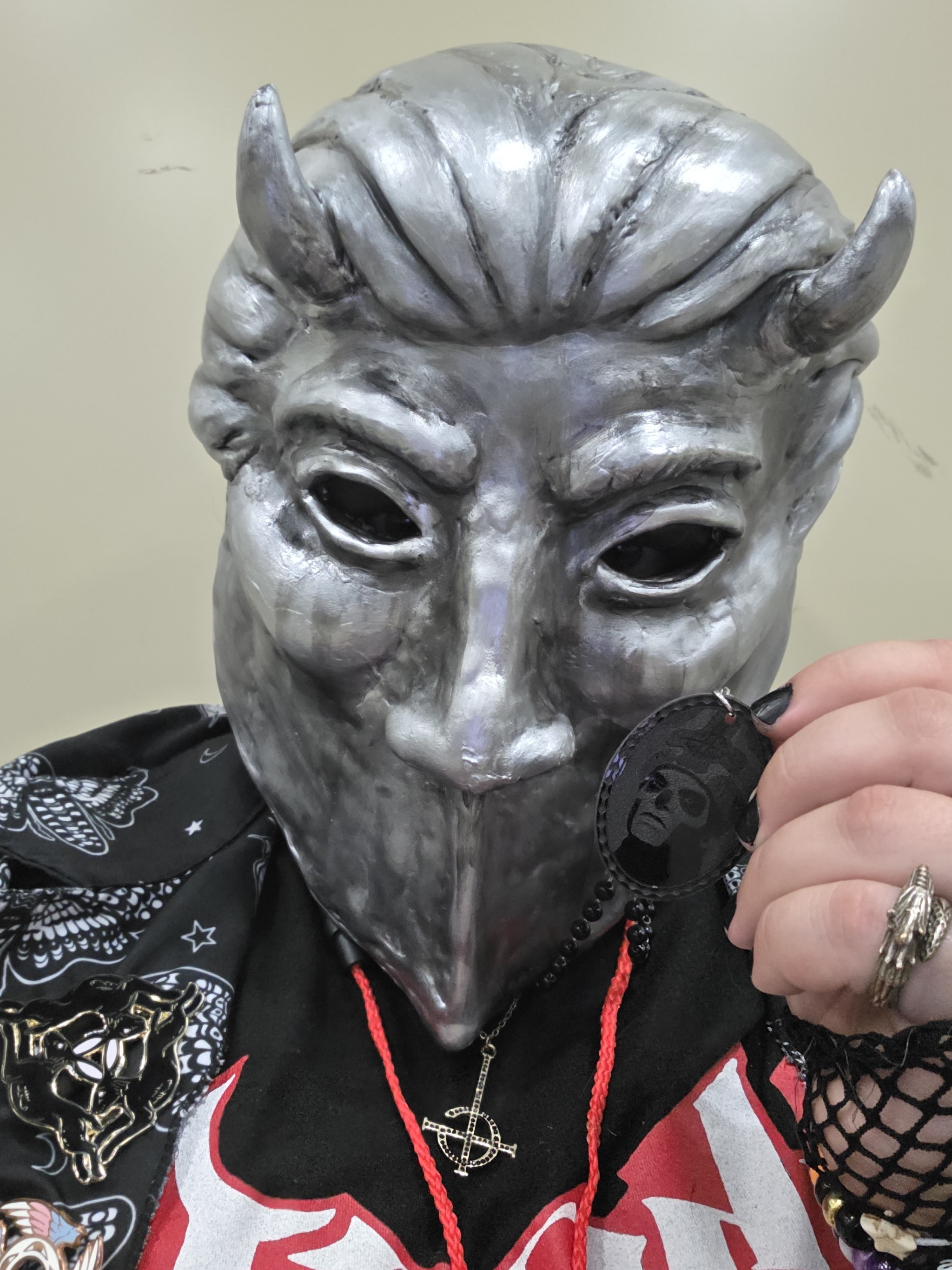 Tiercel Green in a Nameless Ghoul Mask holds up a small plastic token attached to his pentacle rosary with an image of Papa Emeritus III/Terzo on it